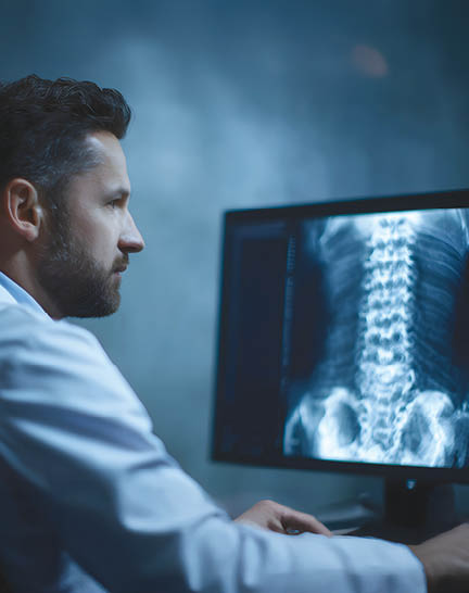 A focused doctor wearing a white lab coat analyzes a detailed X ray image of a patient s spinal column on a bright computer screen in a dimly lit medical office